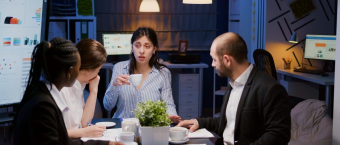 Businesswoman holding cup of coffee while discussing with multi-ethnic teamwork solving management project using graphs paperwork. Diverse coworkers working in meeting room late at night